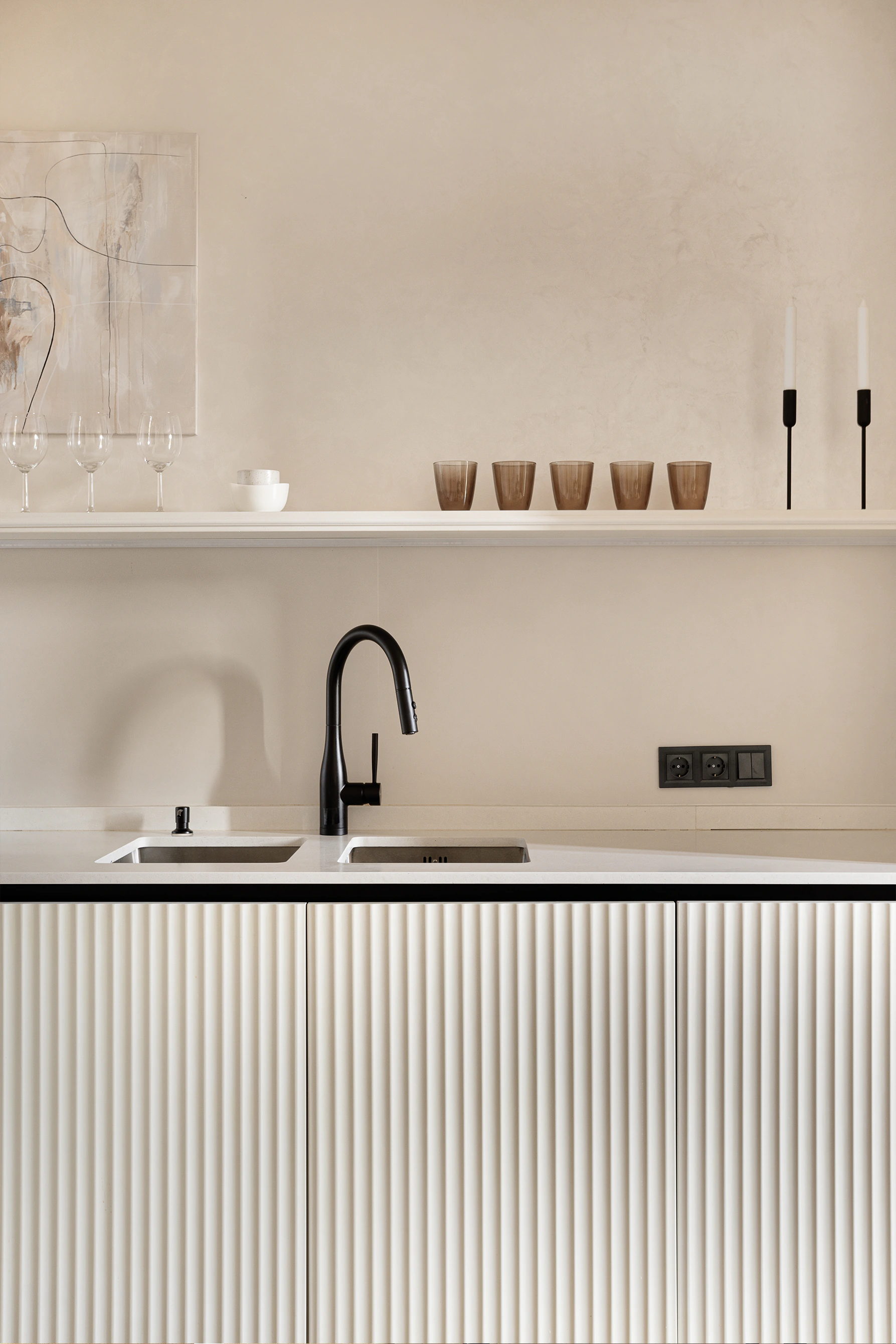 Arcus finished kitchen—double sink with black faucet, fluted cream cabinets, open shelf with glasses and cups, abstract art on plaster wall