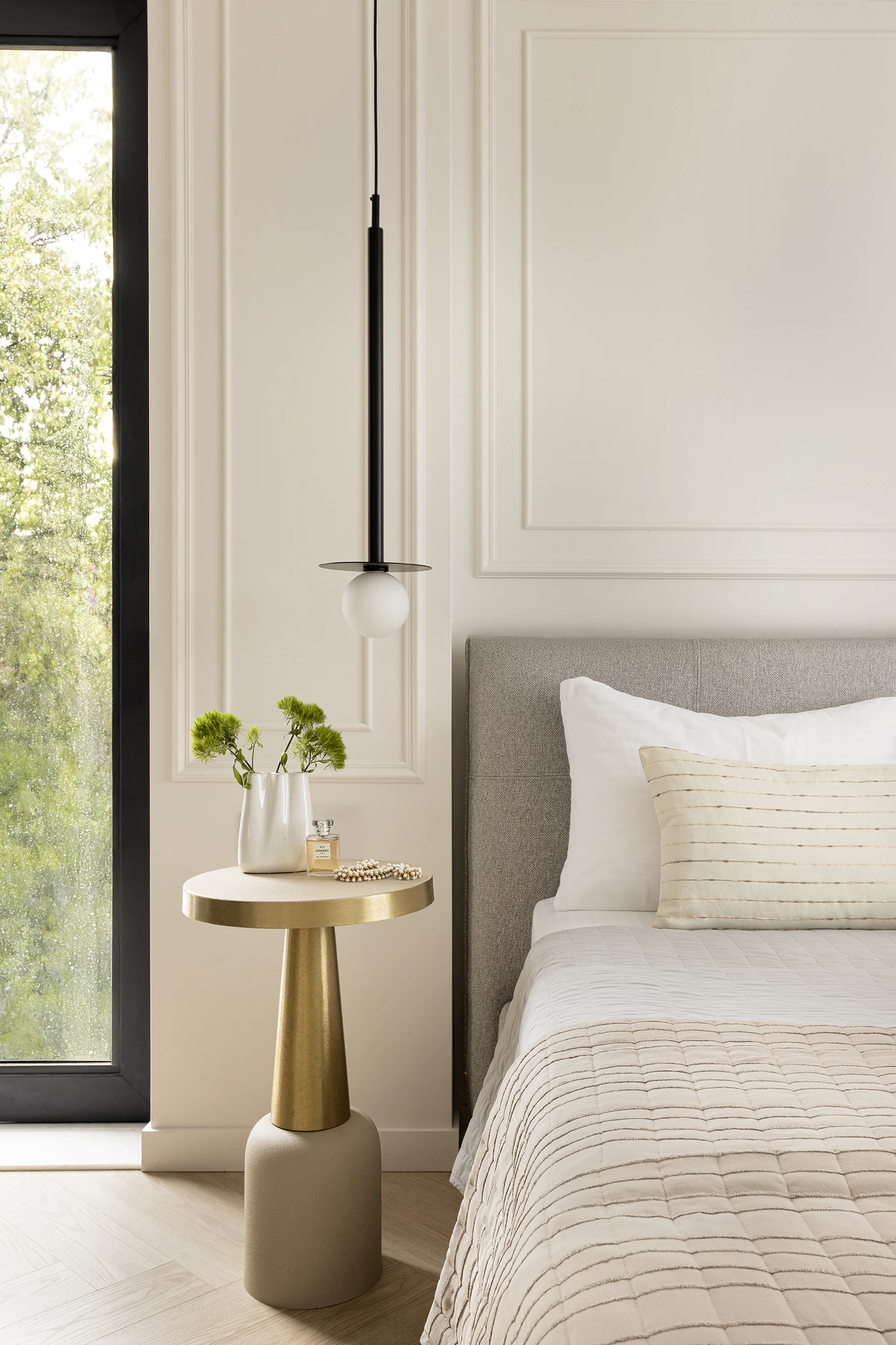 Arcus finished bedroom detail—gold pedestal side table with flowers and perfume, pendant light, grey upholstered headboard
