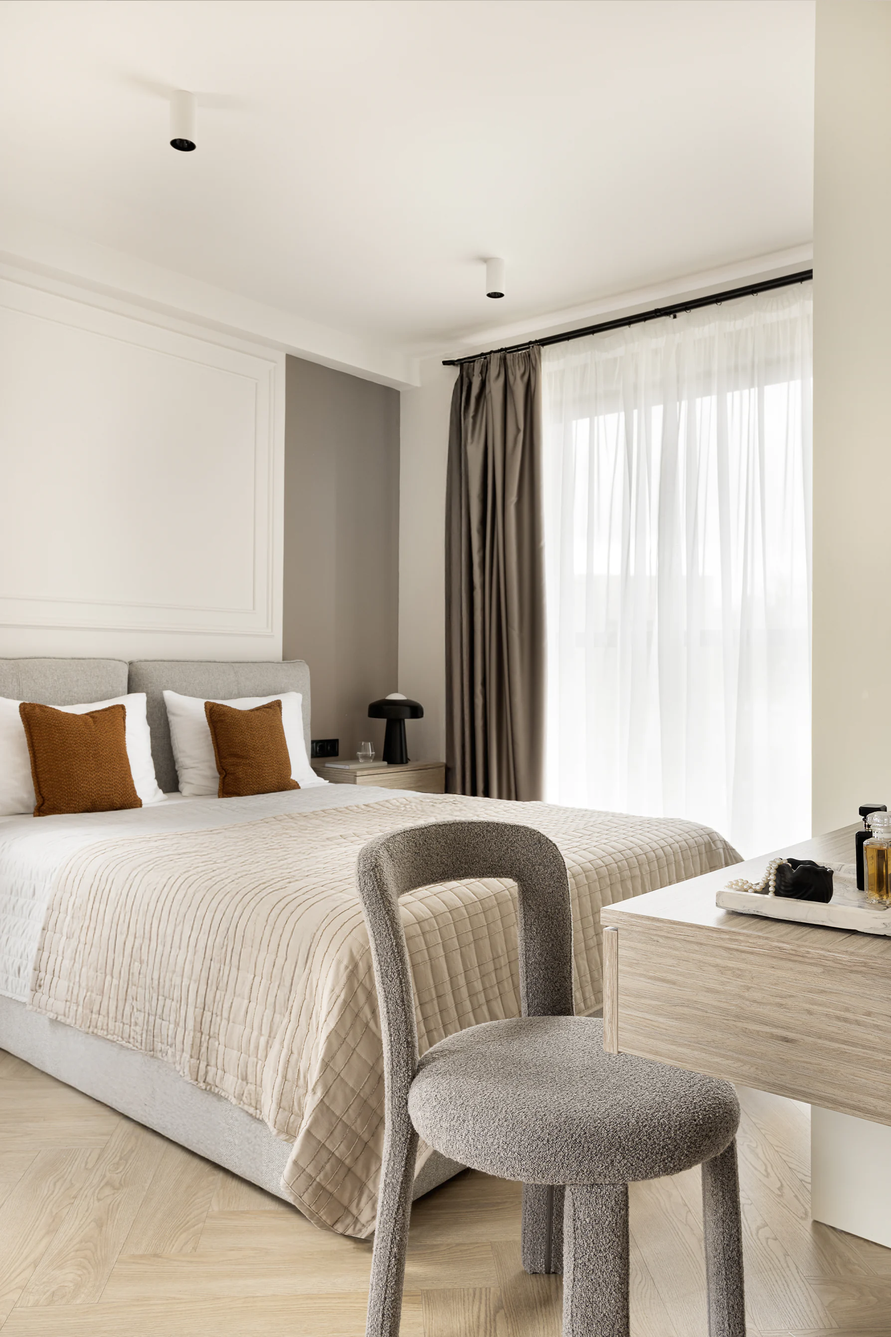 Bedroom—upholstered bed with rust pillows and quilted cover, boucle chair at vanity, sheer curtains, light wood herringbone floor