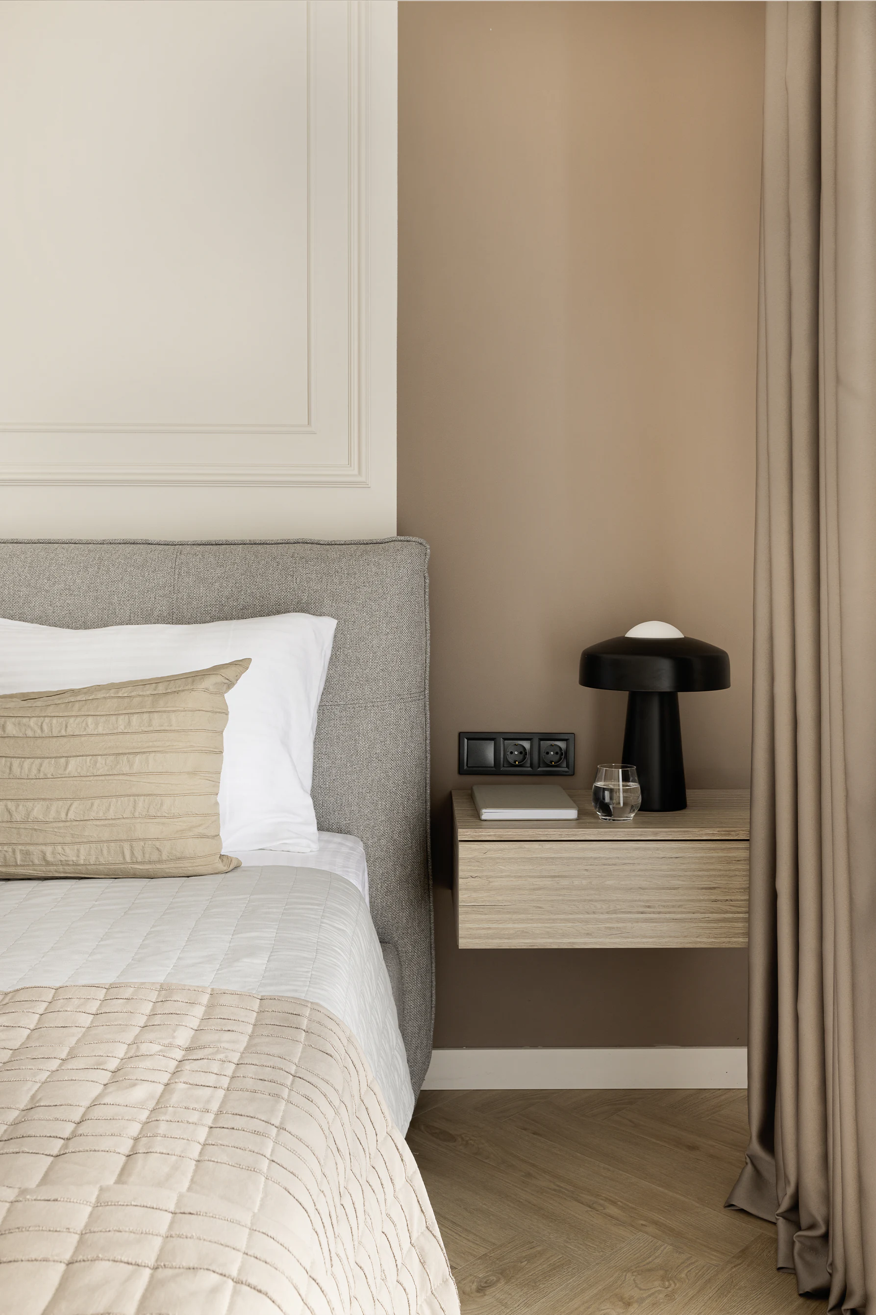 Bedroom detail—grey upholstered headboard, white and beige bedding, floating wood nightstand with black lamp and glass, taupe curtains