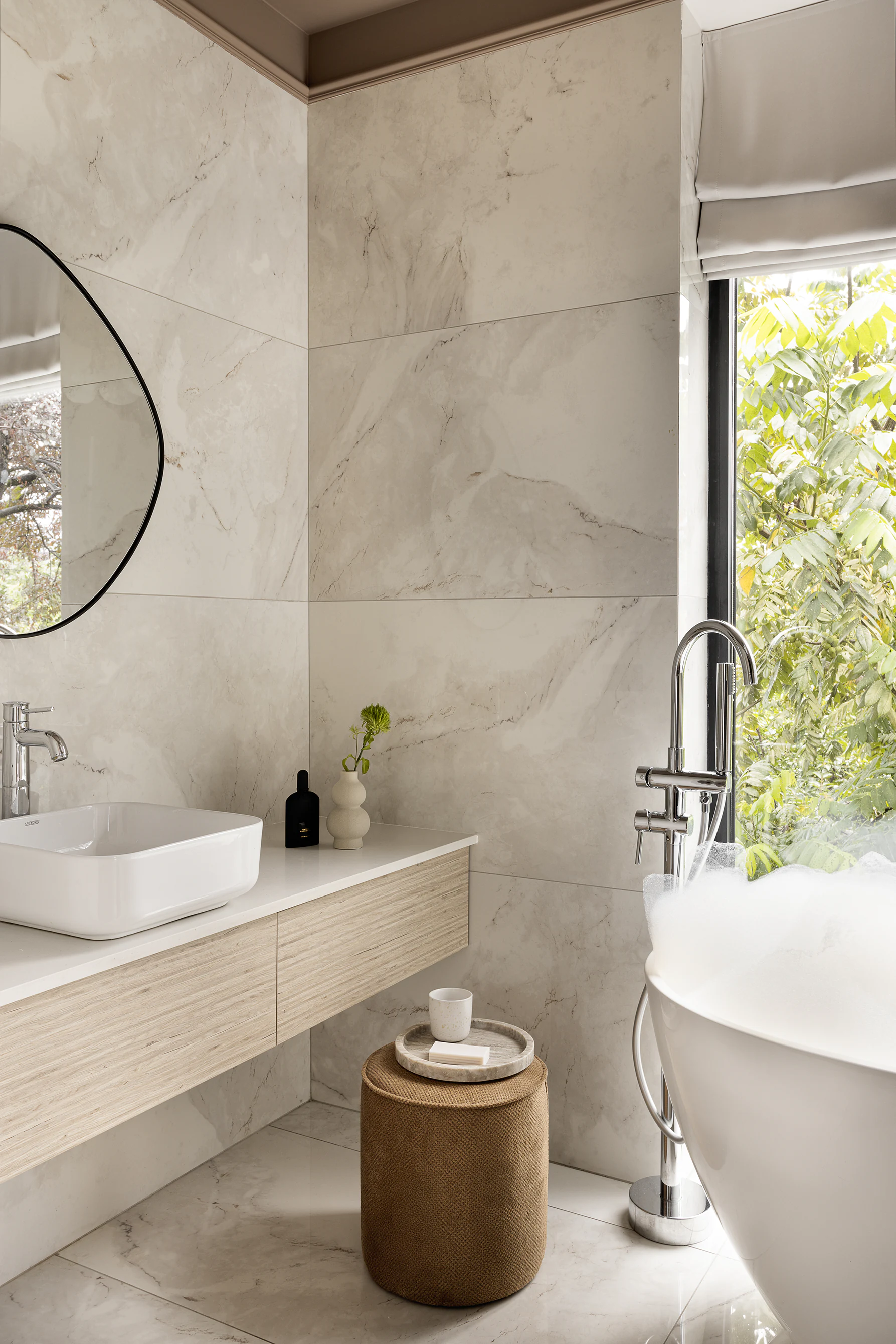 Bathroom vanity—floating wood cabinet with white vessel sink and chrome faucet, round mirror, window to greenery, ottoman with tray