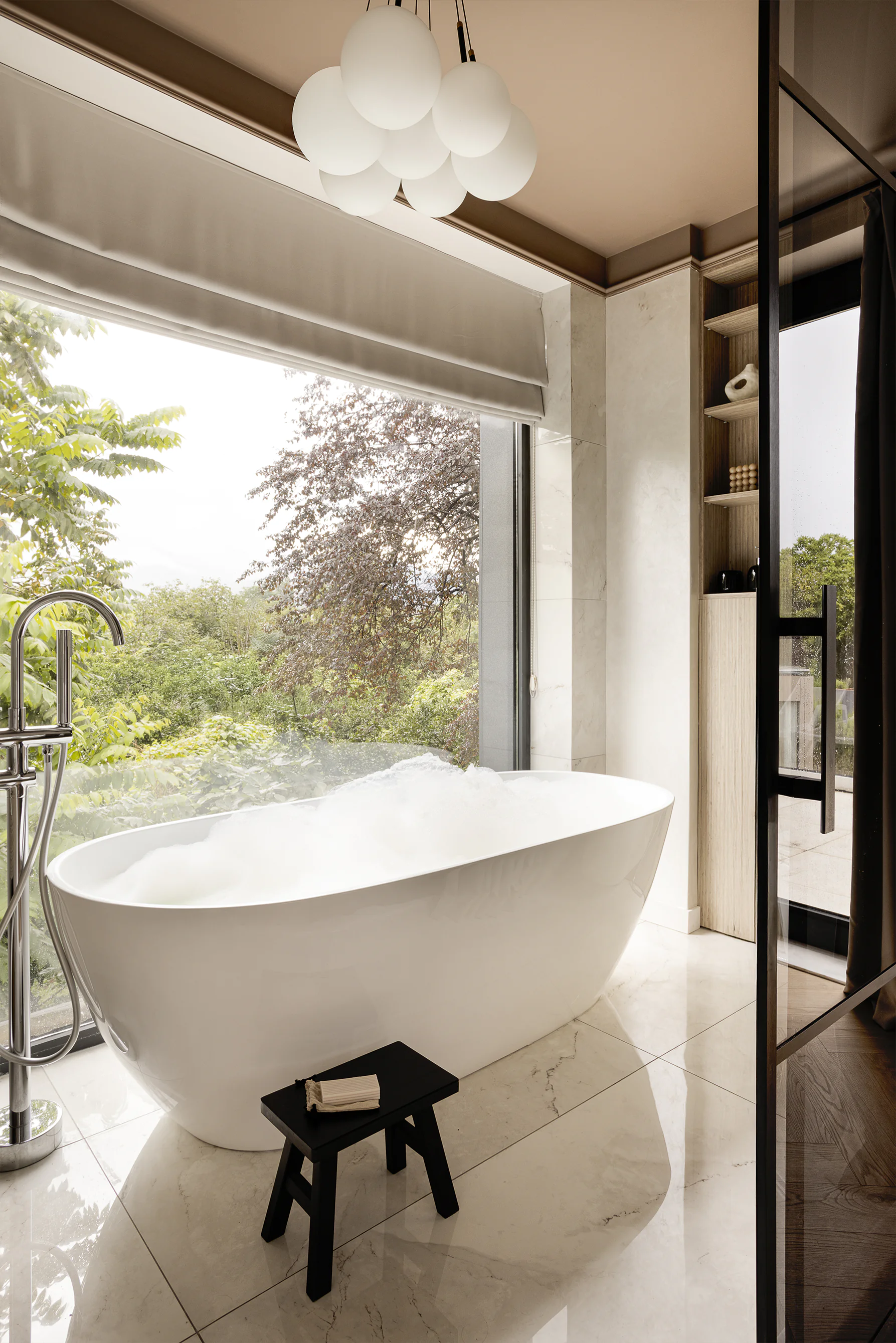 Bathroom with freestanding tub by tall window facing greenery, globe pendant, marble floor, small black stool