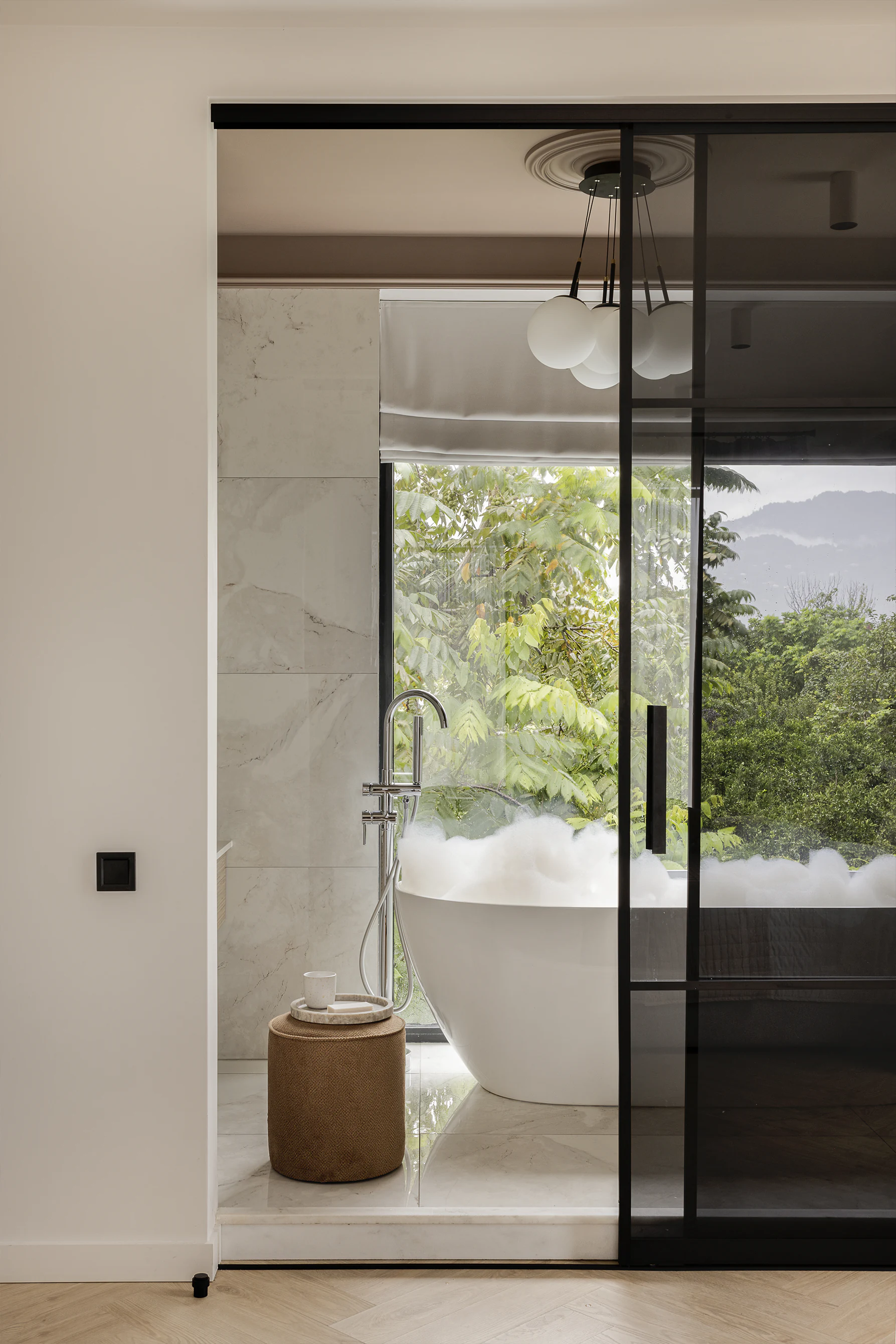 Bathroom—freestanding tub with foam by tall window facing greenery and hills, smoked glass sliding door, ottoman with tray