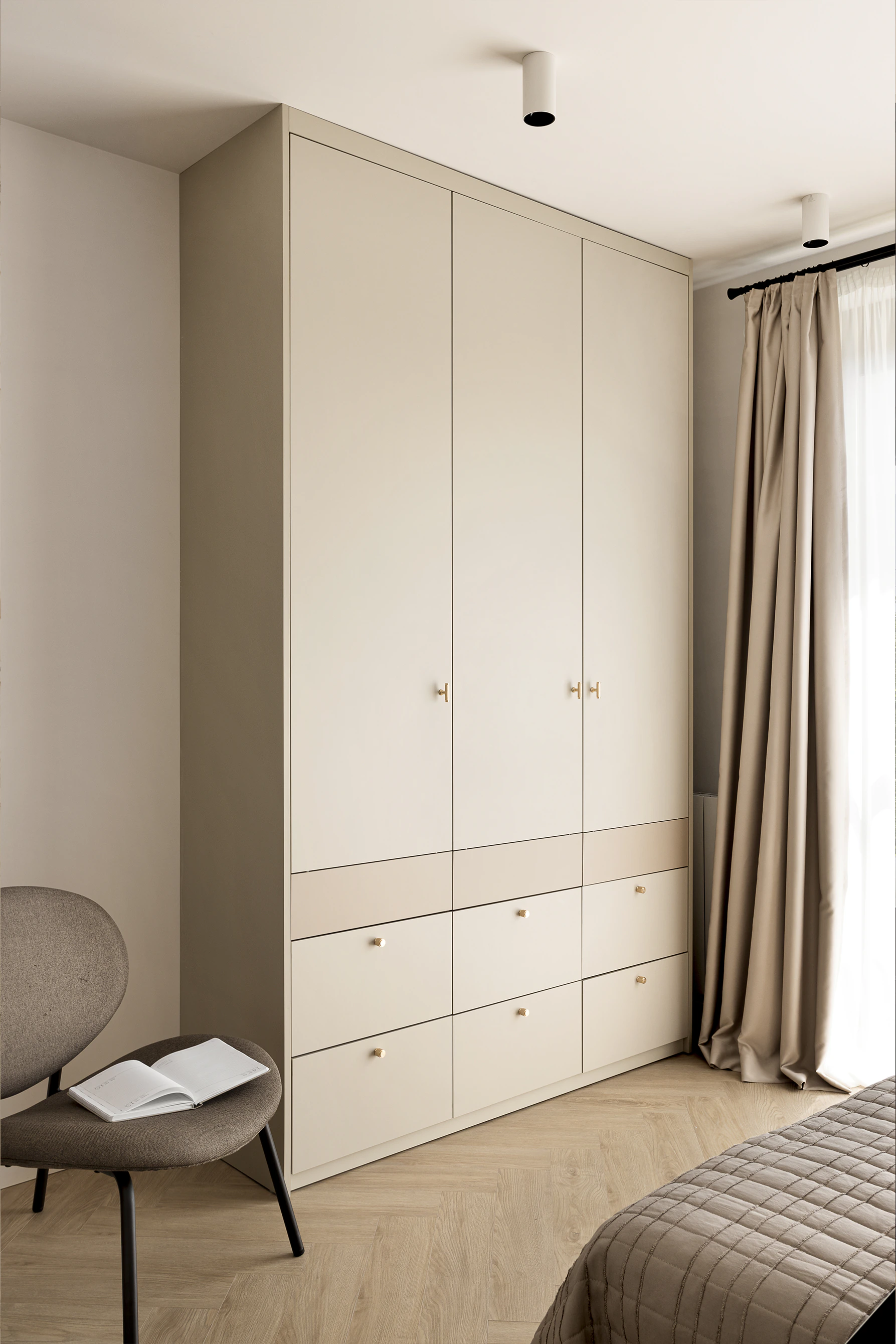 Bedroom—built-in cream wardrobe with drawers, taupe curtains, boucle chair with open book, light herringbone floor