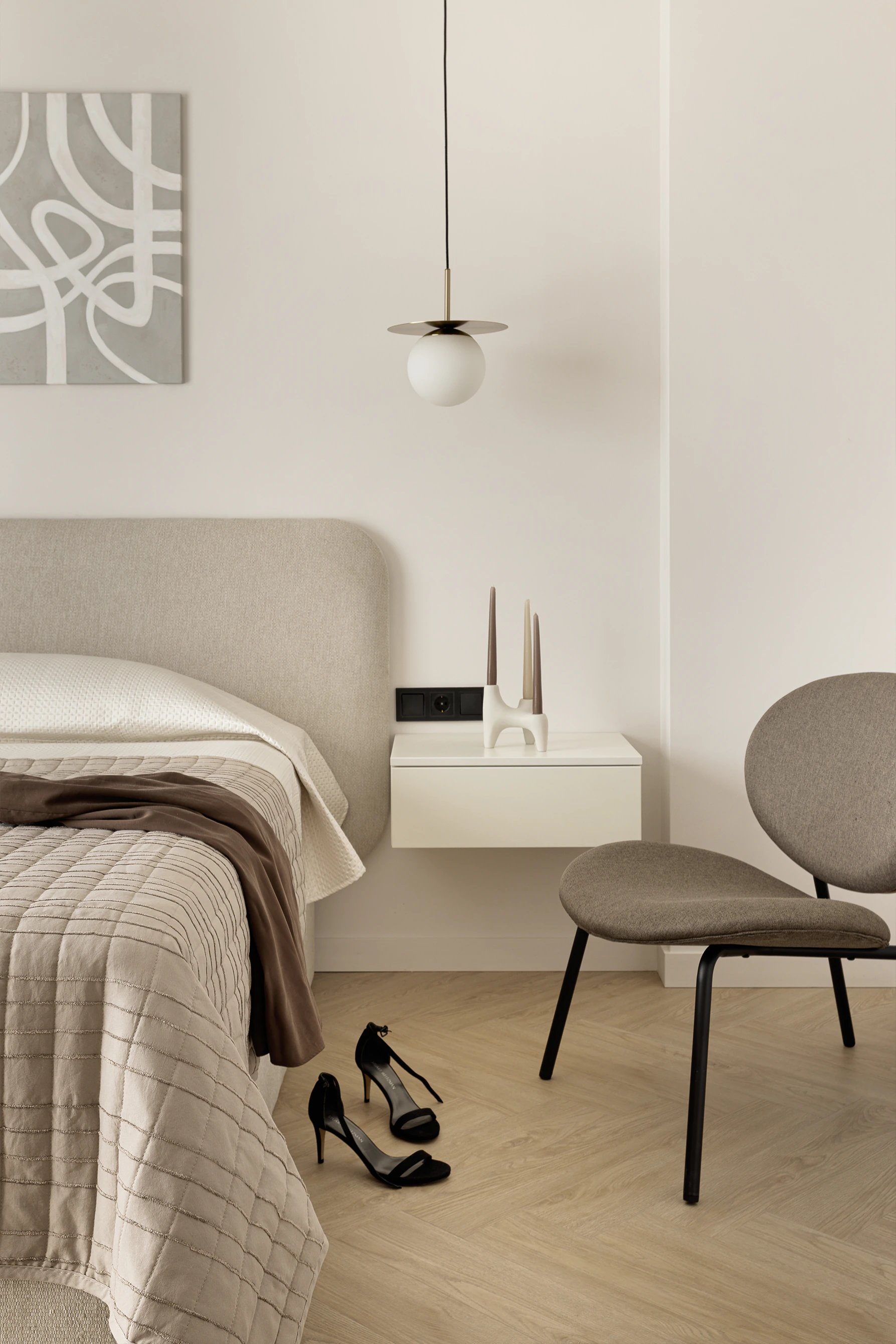Bedroom detail—upholstered headboard, floating nightstand with sculptural candle holder, lounge chair, black heels on floor