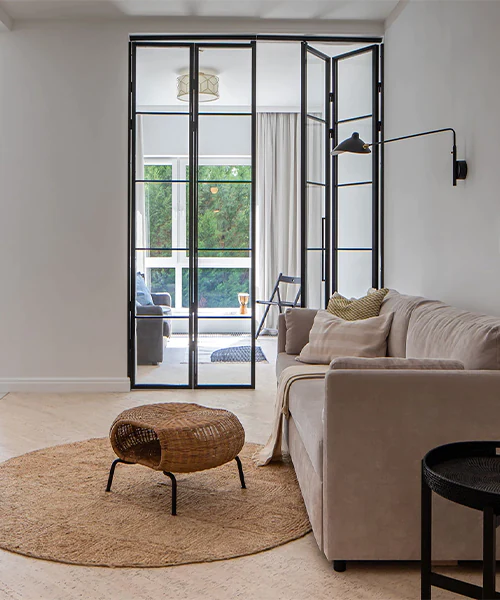Modern living room with a beige sofa, woven coffee table, wall lamp, and black-framed glass partition doors leading to a bright sunroom