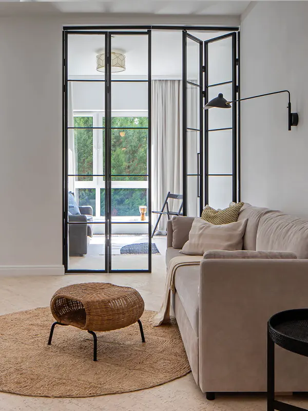Minimalist living room with a beige sofa, woven coffee table, black-framed glass doors, and soft natural light